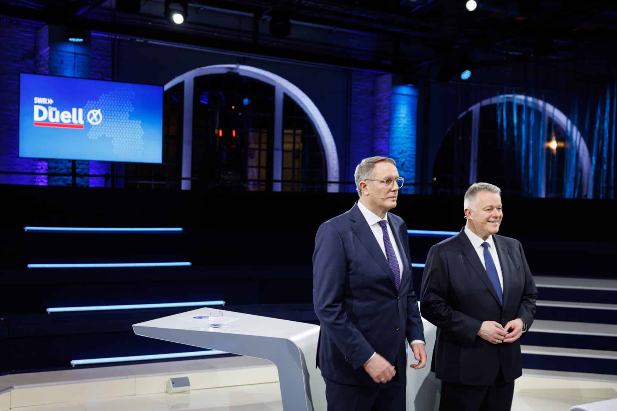 Zwei Politiker stehen im Studio vor einem moderneren Bühnenhintergrund, bereit für eine Fernsehsendung.