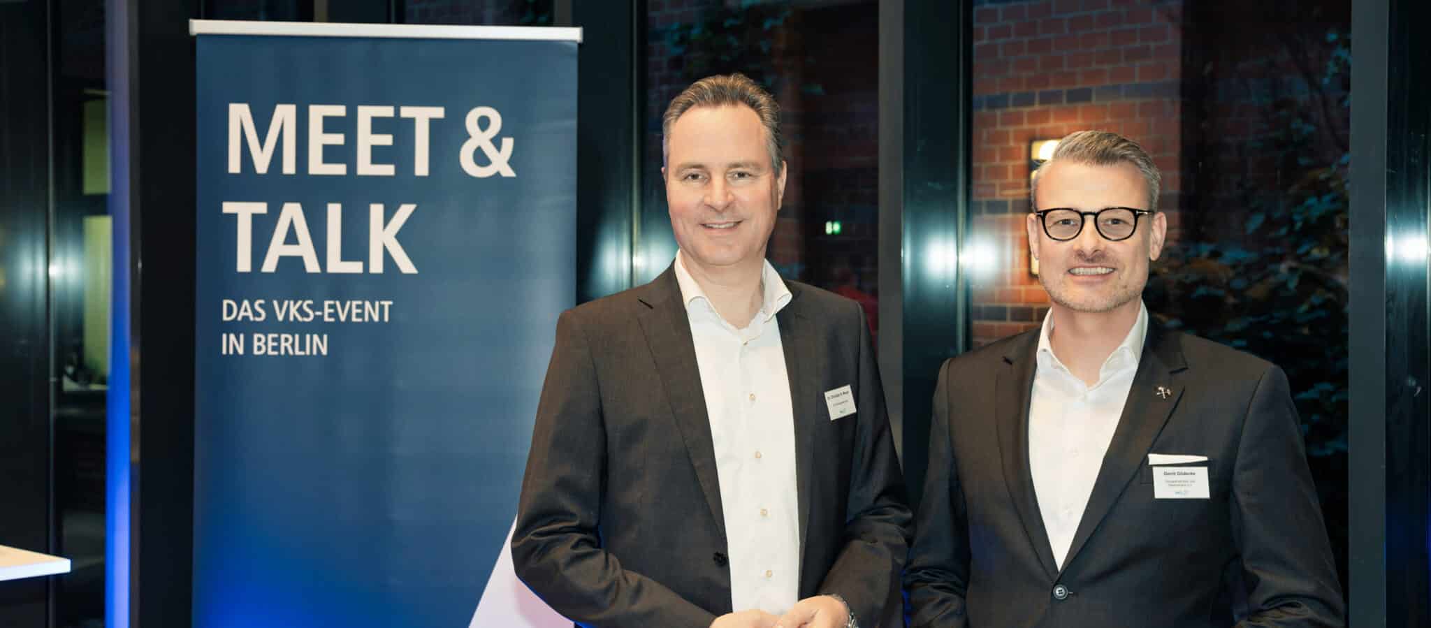 Zwei Männer in Anzügen stehen vor einem Banner mit der Aufschrift "MEET & TALK" bei einem VKS-Event in Berlin.