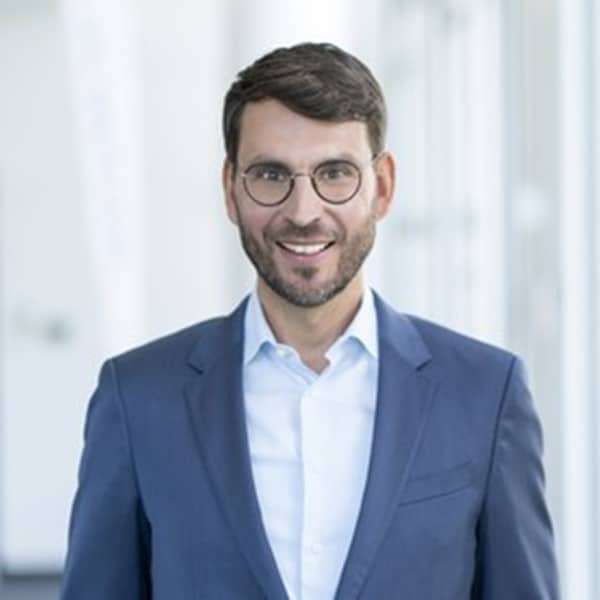 Mann mit Brille und Anzug lächelt in einem modernen Büro mit hellem Hintergrund.