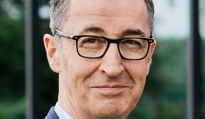 Mann mit Brille und Anzug lächelt in die Kamera, Hintergrund unscharf mit grünen Pflanzen.