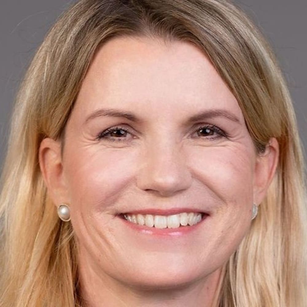 Porträt einer lächelnden Frau mit langen blonden Haaren und Perlenohrringen vor einem neutralen Hintergrund.