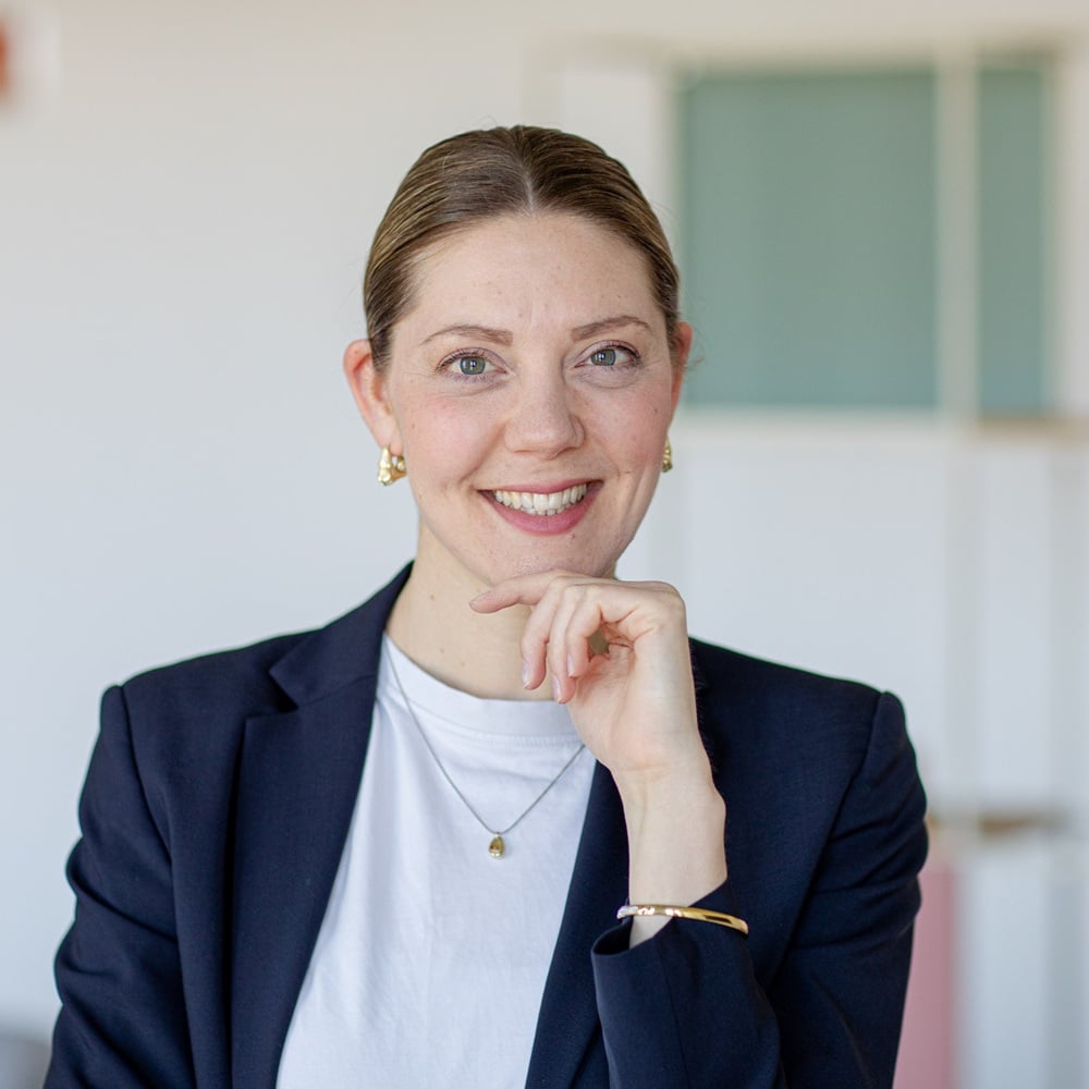 Frau mit kurzem Haar, in Blazer und T-Shirt, lächelt und stützt ihr Kinn mit der Hand in einem modernen Innenraum.