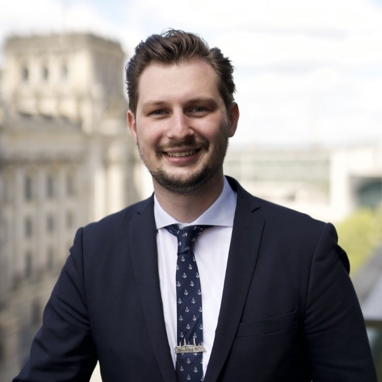 Mann in Anzug mit Krawatte und Anstecknadel lächelt vor urbanem Hintergrund.