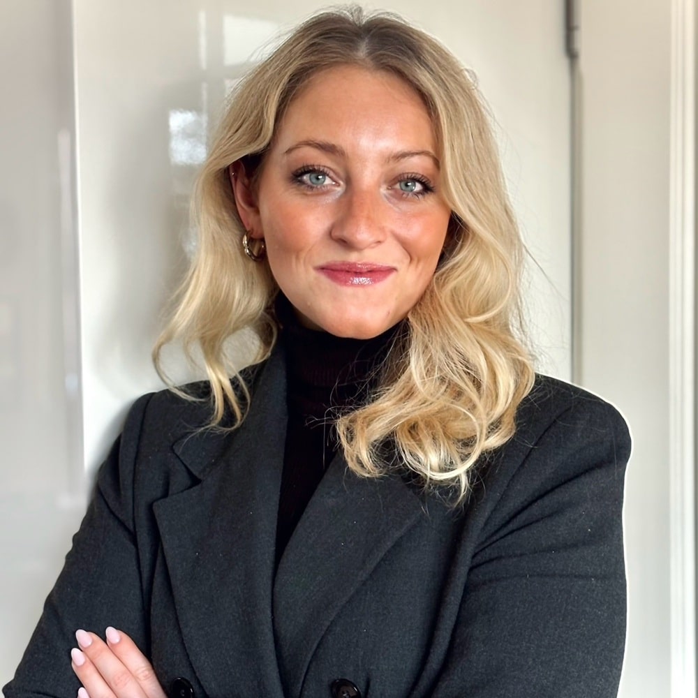 Frau mit blonden, lockigen Haaren und einem schwarzen Blazer, lächelt und steht mit verschränkten Armen vor einer hellen...