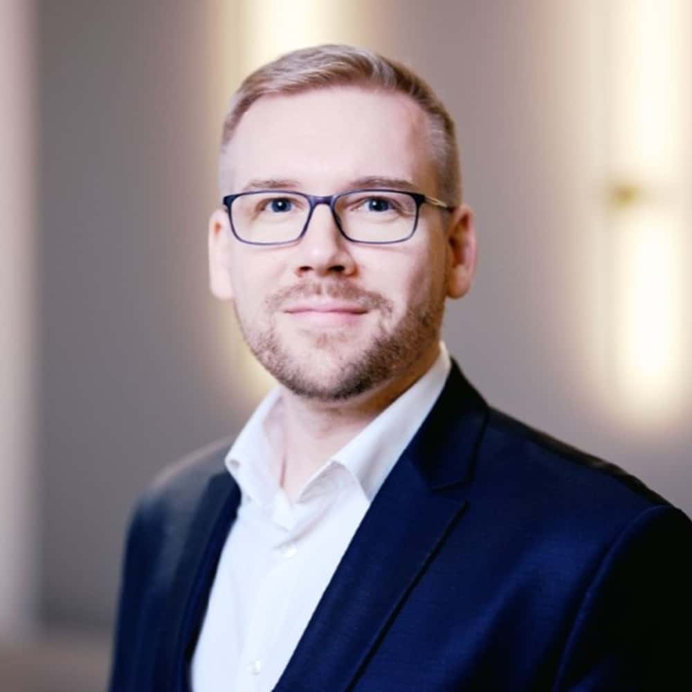 Porträt eines Mannes mit Brille, kurzen blonden Haaren, in einem dunklen Anzug und weißem Hemd, lächelnd vor einem hell...