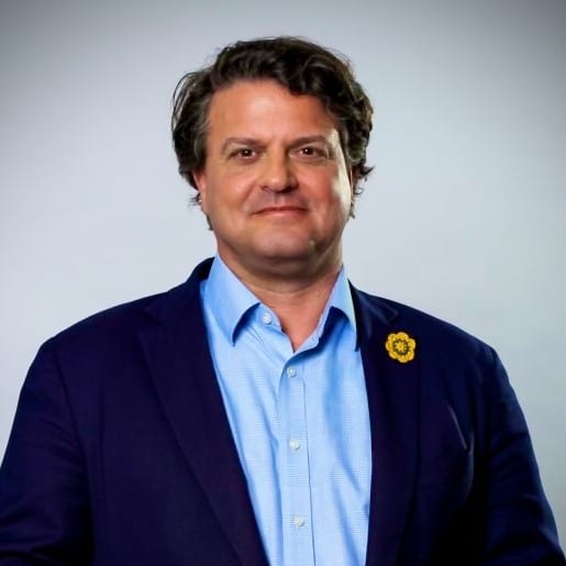 Mann mit lockigem Haar, Anzug und blauer Hemd, lächelt vor neutralem Hintergrund.