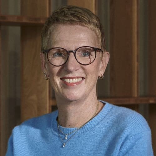 Frau mit kurzen blonden Haaren und Brille, lächelt in einem hellblauen Pullover vor einem Holzregal.