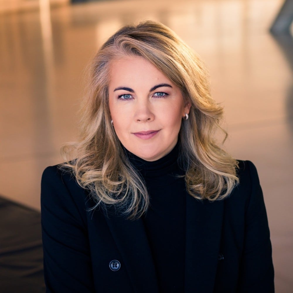 Porträt einer Frau mit blonden, lockigen Haaren, die in einem schwarzen Blazer vor einem modernen Hintergrund sitzt.