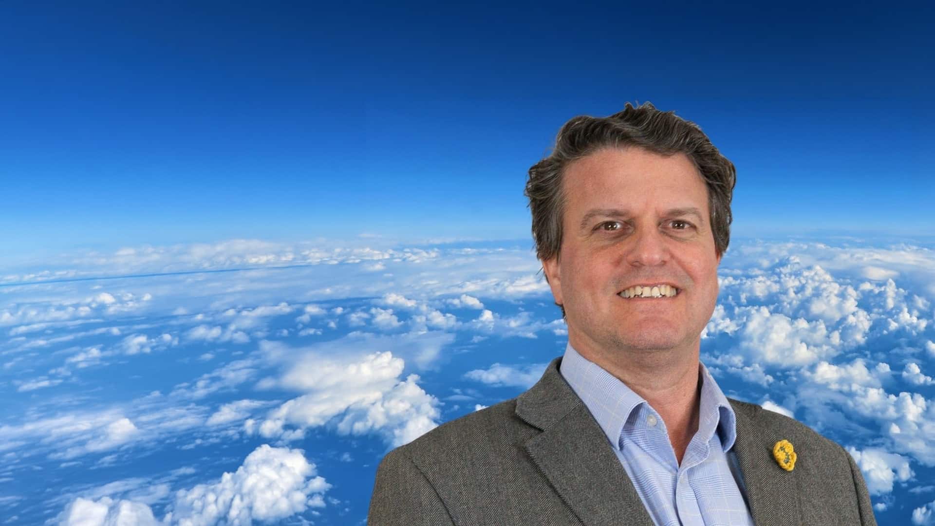Mann mit kurzem Haar, in Anzug, lächelt vor einem Himmel mit Wolken.