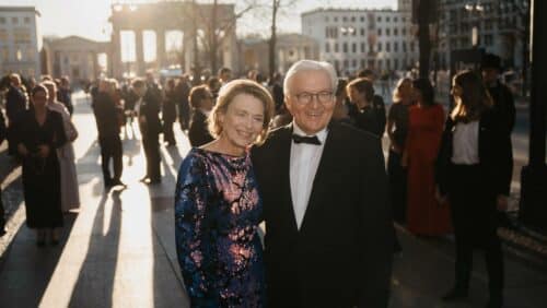 Paar in eleganter Abendkleidung lächelt vor dem Brandenburger Tor, umgeben von festlich gekleideten Menschen im Sonnenlicht.