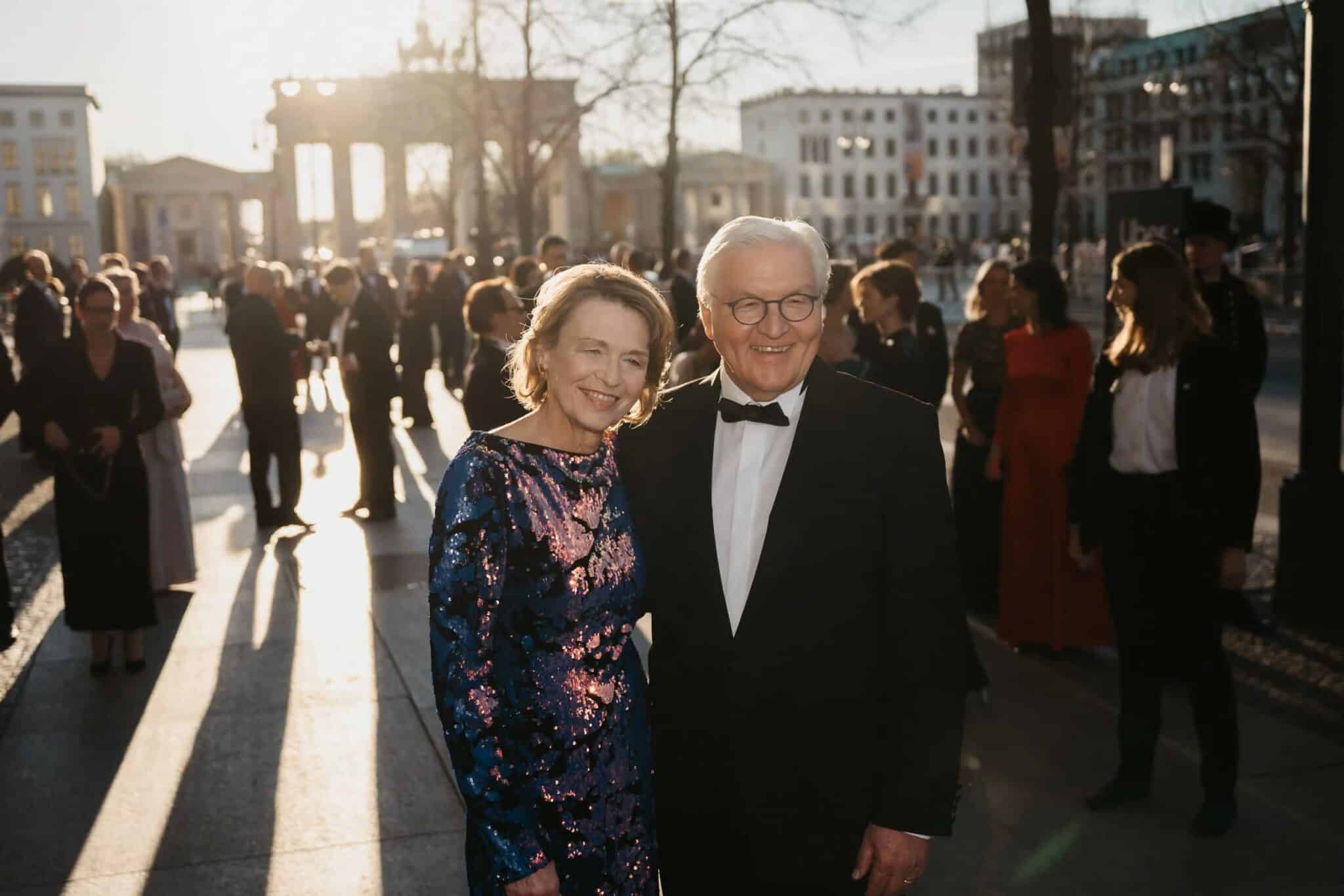 Paar in eleganter Abendkleidung lächelt vor dem Brandenburger Tor, umgeben von festlich gekleideten Menschen im Sonnenlicht.