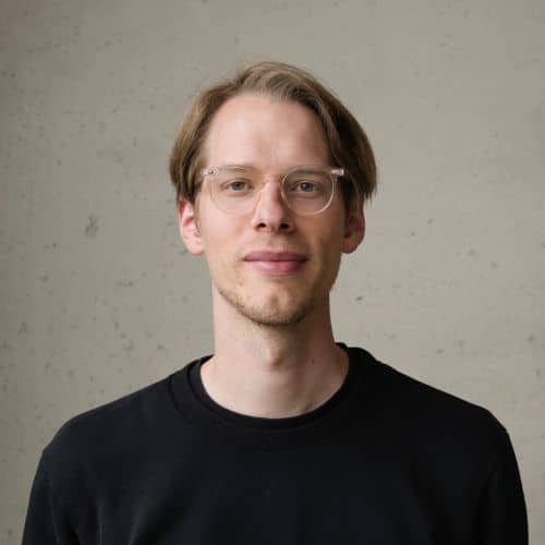 Junger Mann mit Brille und blonden Haaren, trägt ein schwarzes Sweatshirt, vor einer grauen Wand.