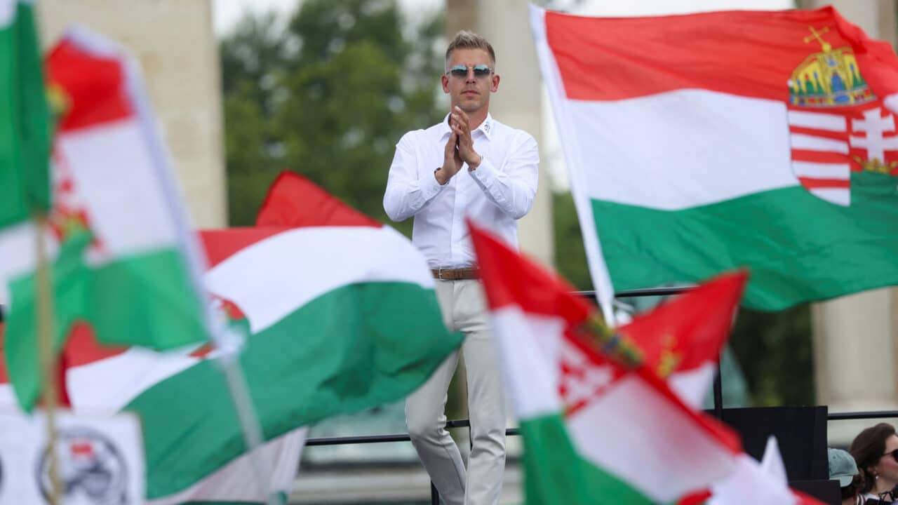 Péter Magyar steht auf einer Bühne, umgeben von ungarischen Flaggen, und applaudiert.
