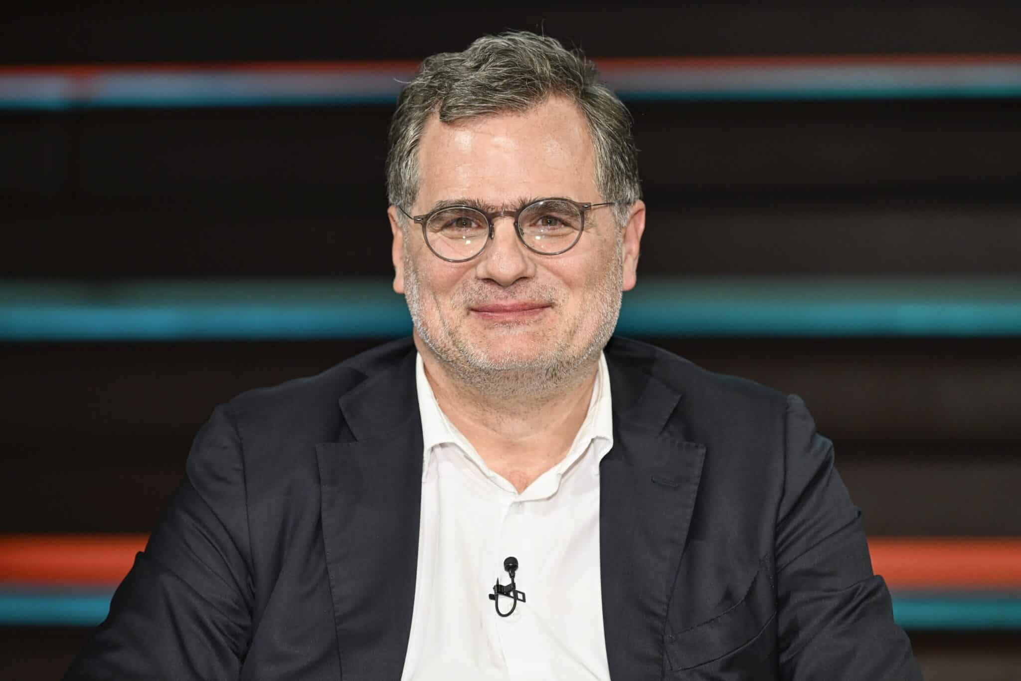 Wolfgang Schmidt, lächelnd, trägt Brille und dunklen Anzug, sitzt in einer Talkshow-Umgebung mit bunten Stufen im Hinter...