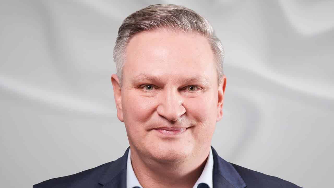 Porträt eines Mannes mit grauen Haaren, lächelnd, in einem dunkelblauen Anzug vor einem hellen, verschwommenen Hintergrund.
