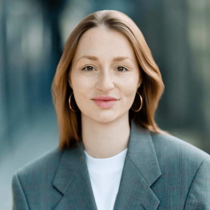 Porträt einer Frau mit langen, braunen Haaren, die einen grauen Blazer trägt und in die Kamera lächelt.