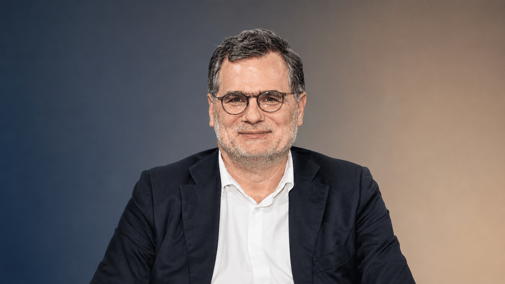 Mann mit Brille und Anzug lächelt vor einem neutralen Hintergrund in sanften Farbtönen.