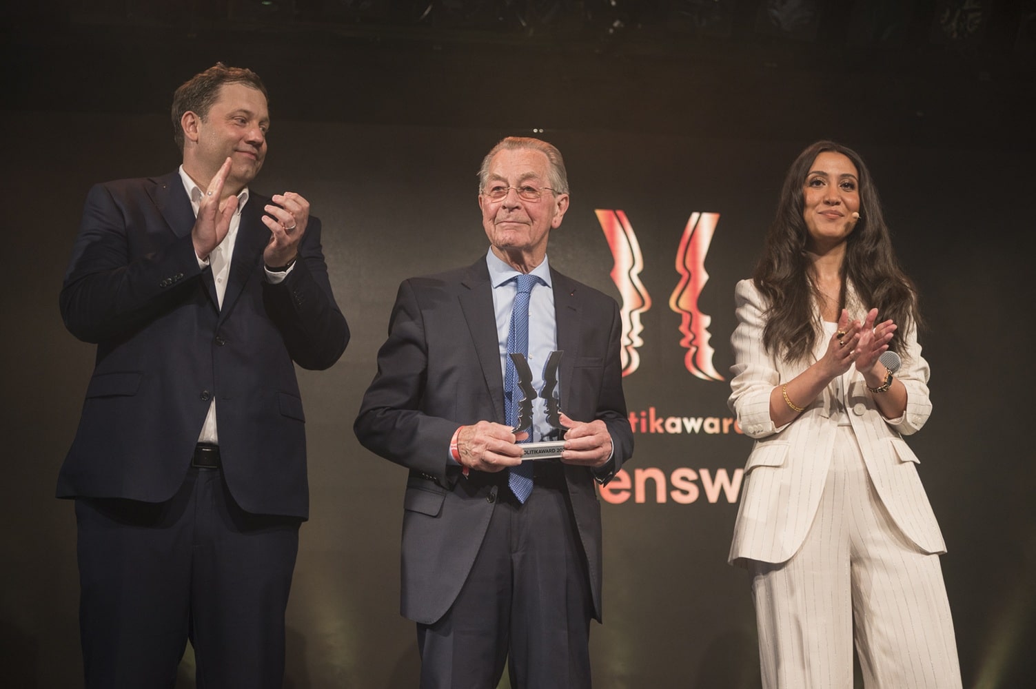 Drei Personen applaudieren auf der Bühne bei der Verleihung des Politikawards 2026. Hintergrund mit Logo des Events.