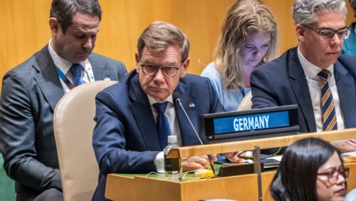 Männer und Frauen sitzen an einem Tisch im UN-Saal, ein Mann mit Brille und Schild "Germany" schaut nachdenklich.