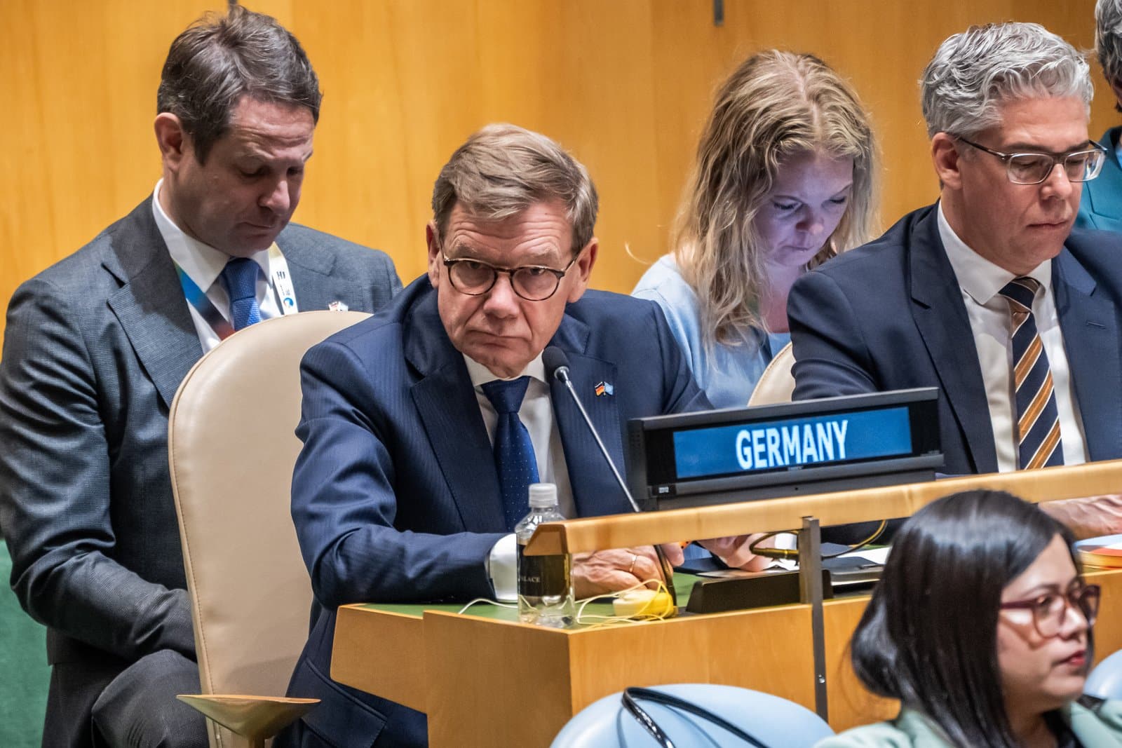 Männer und Frauen sitzen an einem Tisch im UN-Saal, ein Mann mit Brille und Schild "Germany" schaut nachdenklich.