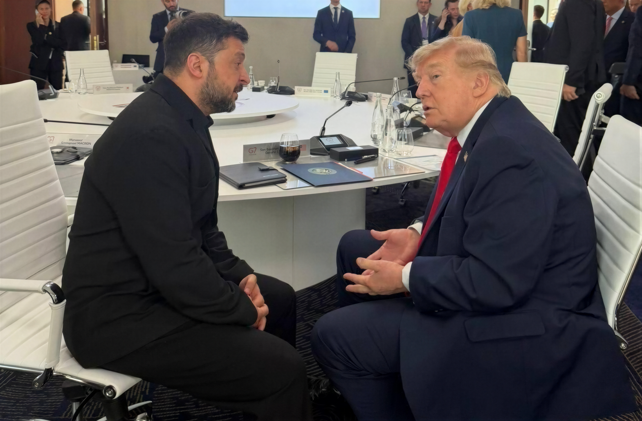 Two men in suits converse at a round conference table, with officials standing in the background.