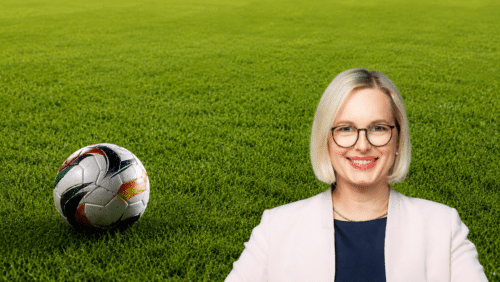 Frau mit blonden, kurzen Haaren und Brille lächelt vor einem Fußball auf grünem Rasen.