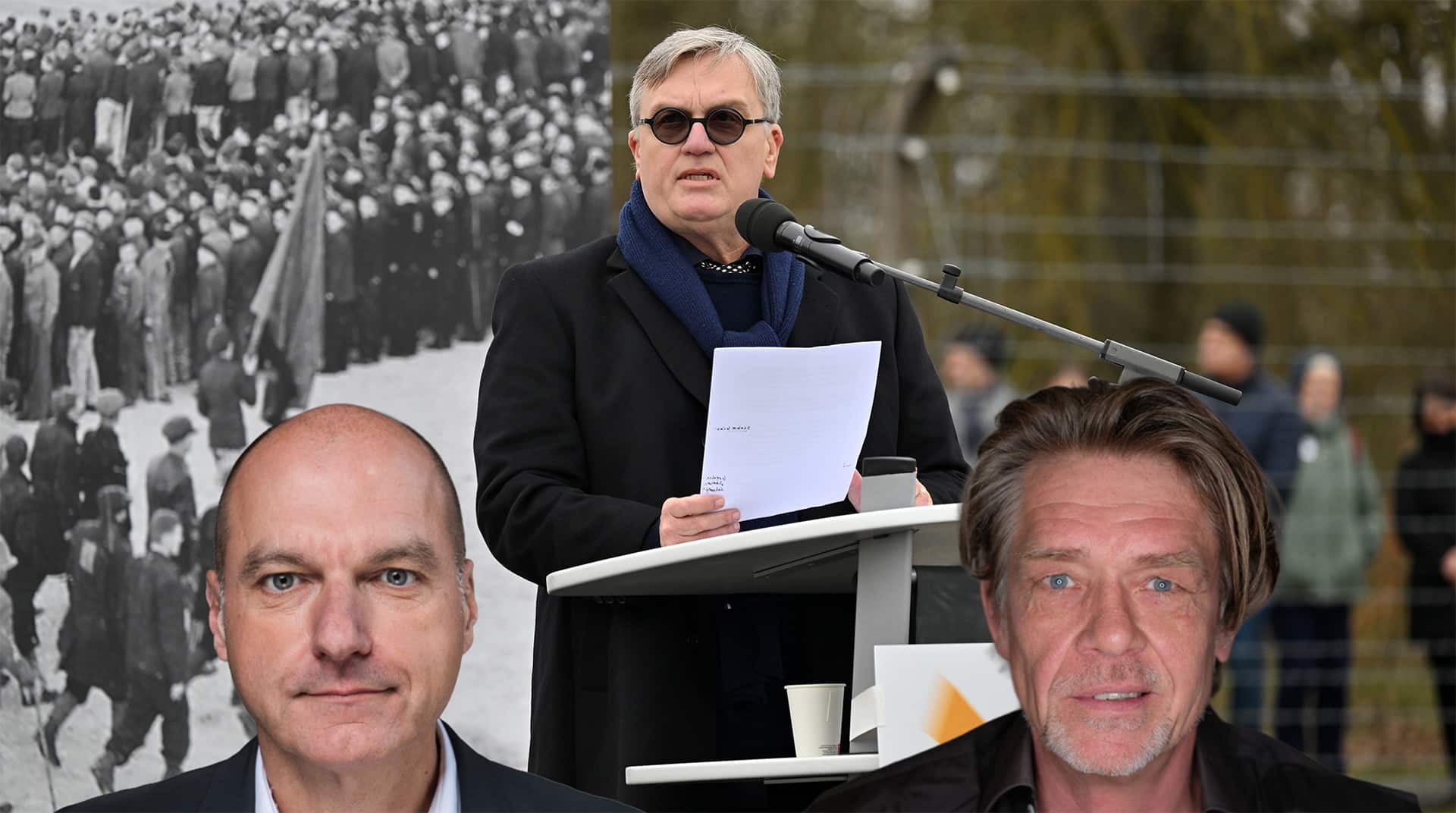 Mann am Mikrofon spricht vor einer Menschenmenge, mit zwei weiteren Männern im Vordergrund.