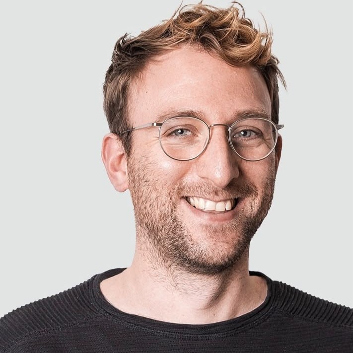Lächelnder Mann mit Brille und lockigem Haar, trägt ein schwarzes Oberteil, vor einem hellen Hintergrund.