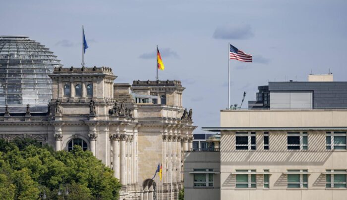 Reichstagsgebäude in Berlin mit deutscher und US-Flagge, umgeben von Bäumen und moderner Architektur im Hintergrund.