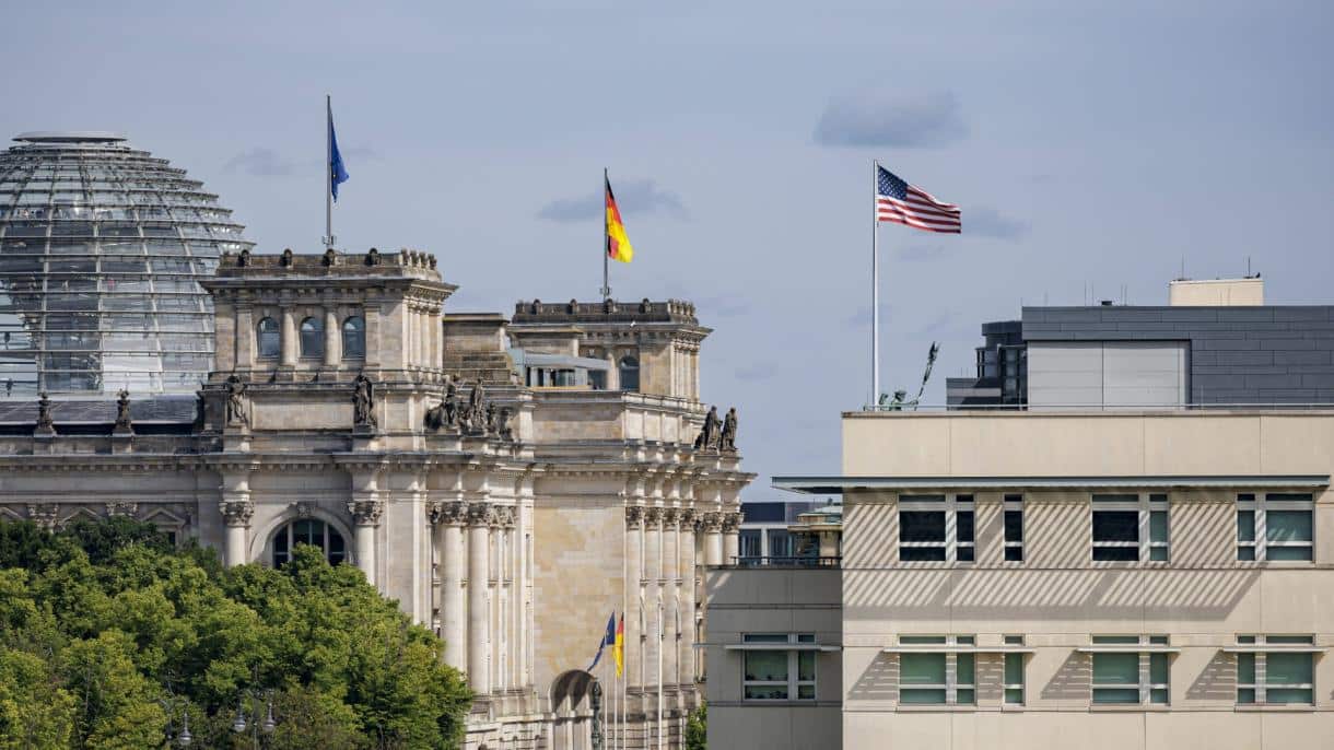 Reichstagsgebäude in Berlin mit deutscher und US-Flagge, umgeben von Bäumen und moderner Architektur im Hintergrund.