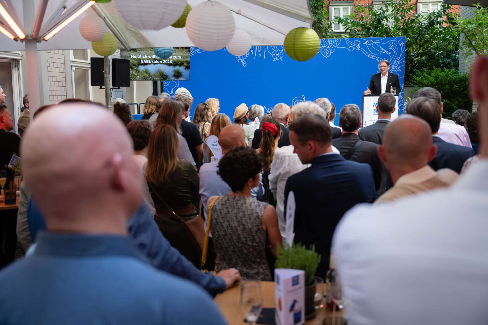 Menschenmenge bei einer Veranstaltung, ein Redner steht auf einer Bühne vor einem blauen Hintergrund mit Pflanzenmotiven.