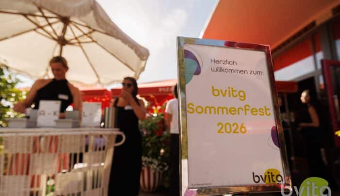 Ein Schild mit der Aufschrift "bvigt Sommerfest 2026" vor einer festlichen Outdoor-Umgebung mit Gästen.