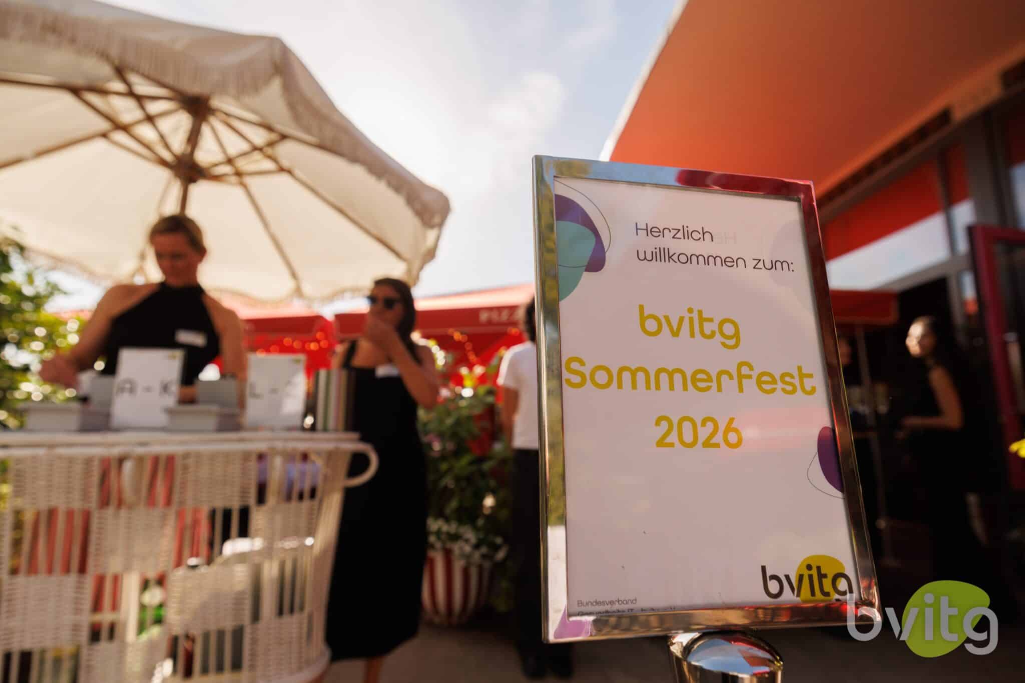 Ein Schild mit der Aufschrift "bvigt Sommerfest 2026" vor einer festlichen Outdoor-Umgebung mit Gästen.