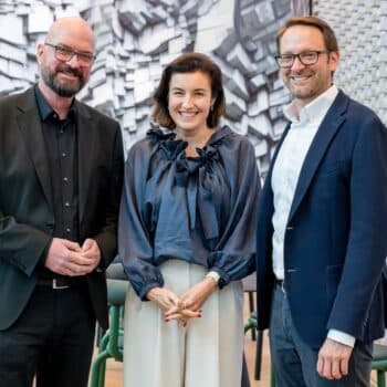 Three professionals pose together in a modern office with textured acoustic wall panels behind them, smiling at the camera.