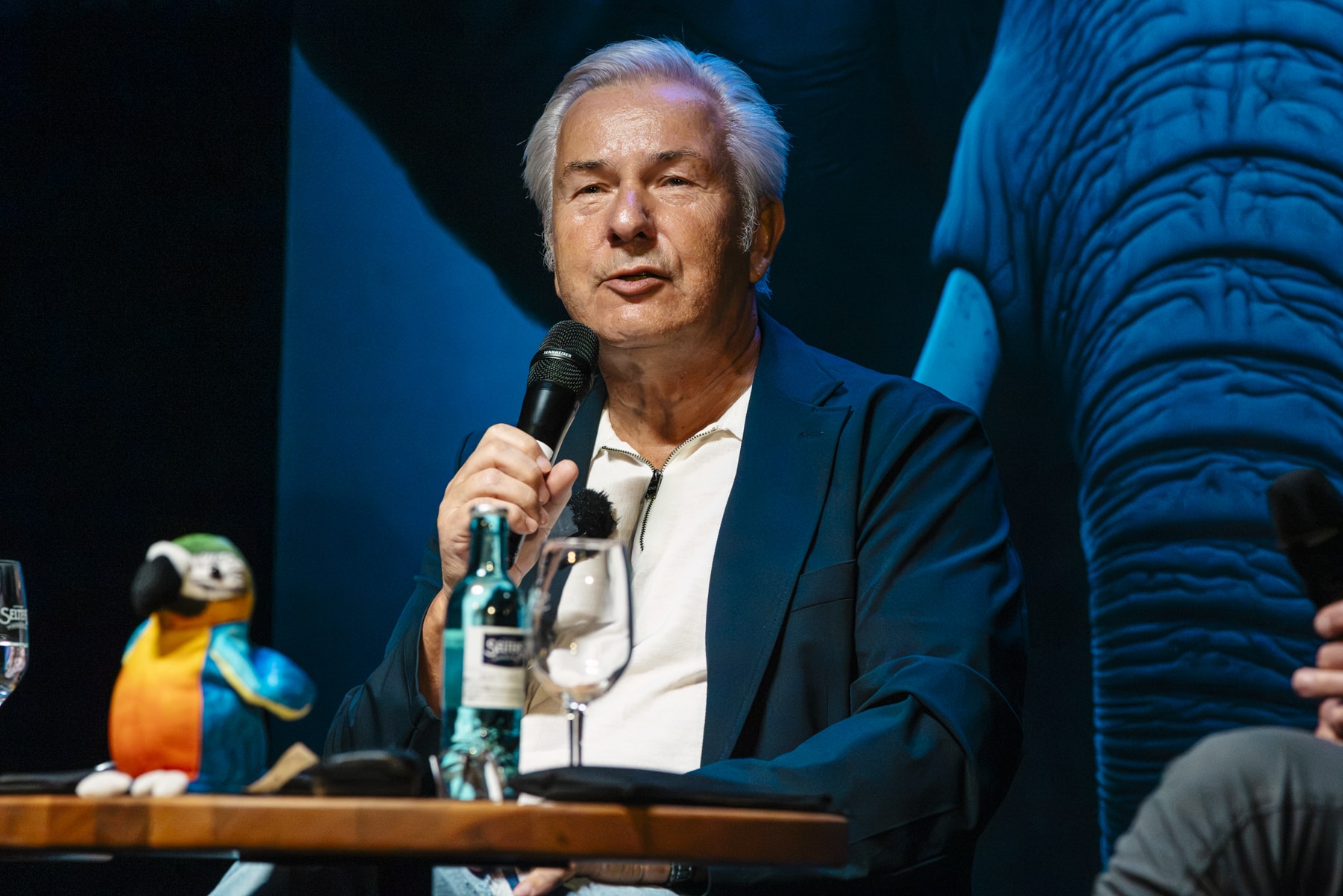 Mann mit Mikrofon spricht, bunte Papageienfigur und Glas Wasser auf Tisch, Elefantenbild im Hintergrund.