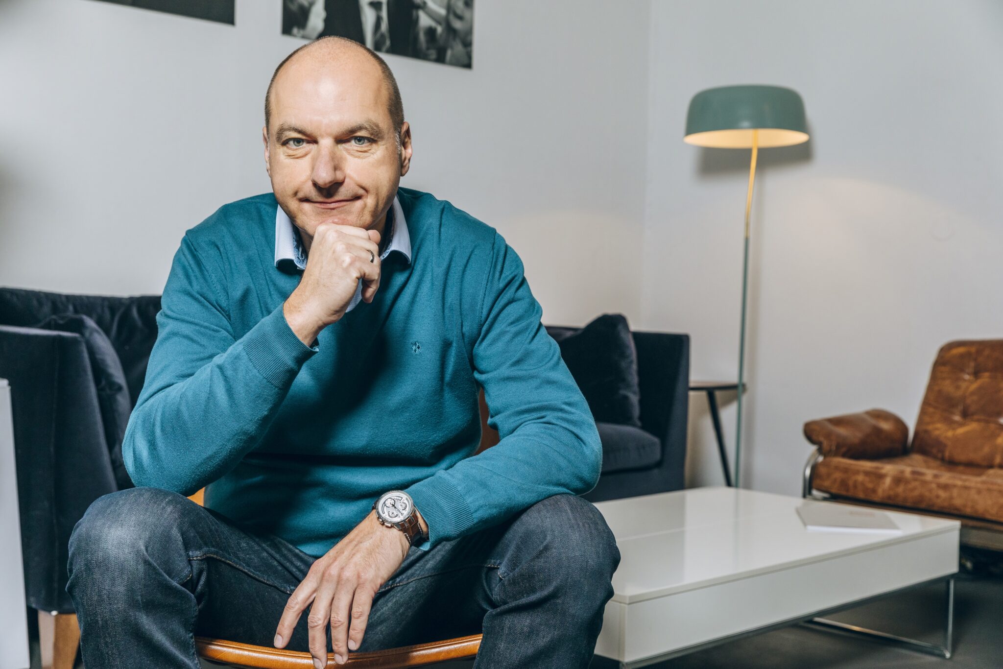 Mann mit kurzem Haar in blauer Pullover sitzt nachdenklich in einem modernen Raum mit Sofa und Lampe.