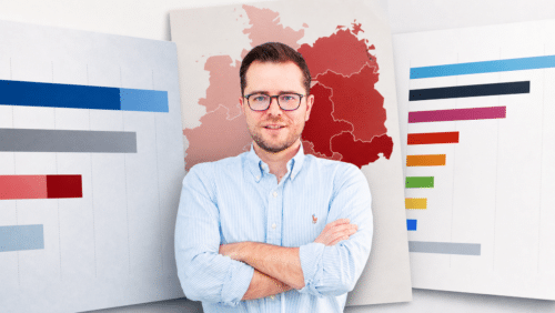 Mann mit Brille und Hemd steht vor Grafiken und Karten, die Daten und Statistiken zu Deutschland zeigen.