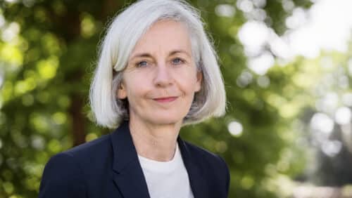 Frau mit grauen Haaren und blauer Jacke steht vor unscharfem, grünem Hintergrund und lächelt leicht.