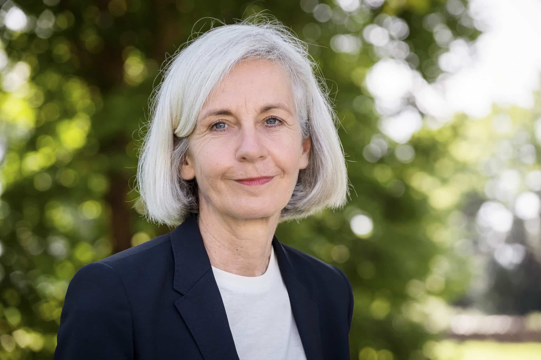 Frau mit grauen Haaren und blauer Jacke steht vor unscharfem, grünem Hintergrund und lächelt leicht.