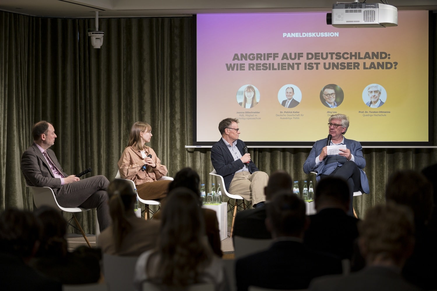 Paneldiskussion mit vier Sprechern auf einer Bühne, im Hintergrund ein buntes Banner mit dem Titel der Veranstaltung.