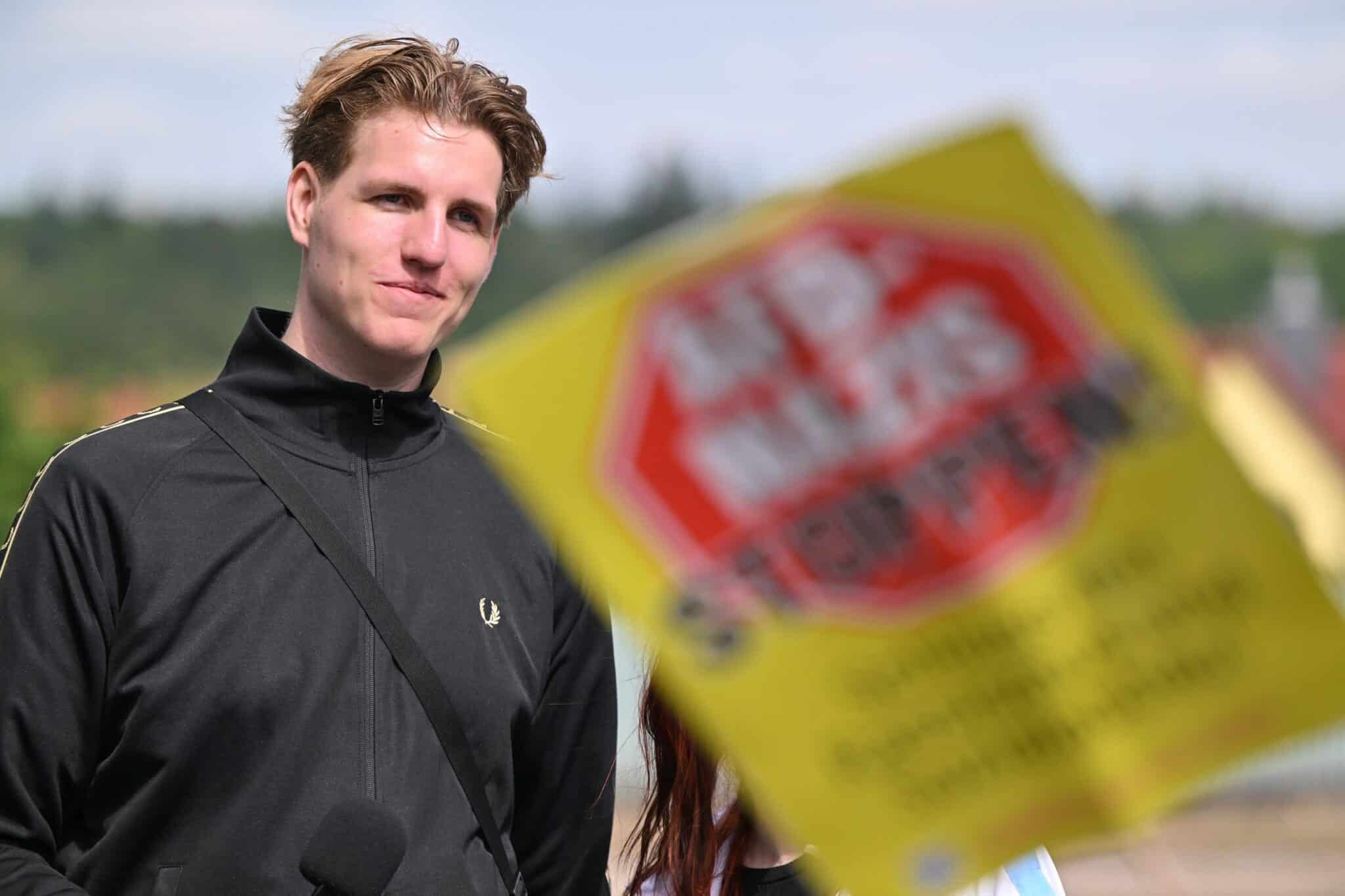 Junger Mann mit schwarzer Jacke steht vor einem unscharfen Protestplakat mit der Aufschrift „AFD-MAXI STOPPEN“.