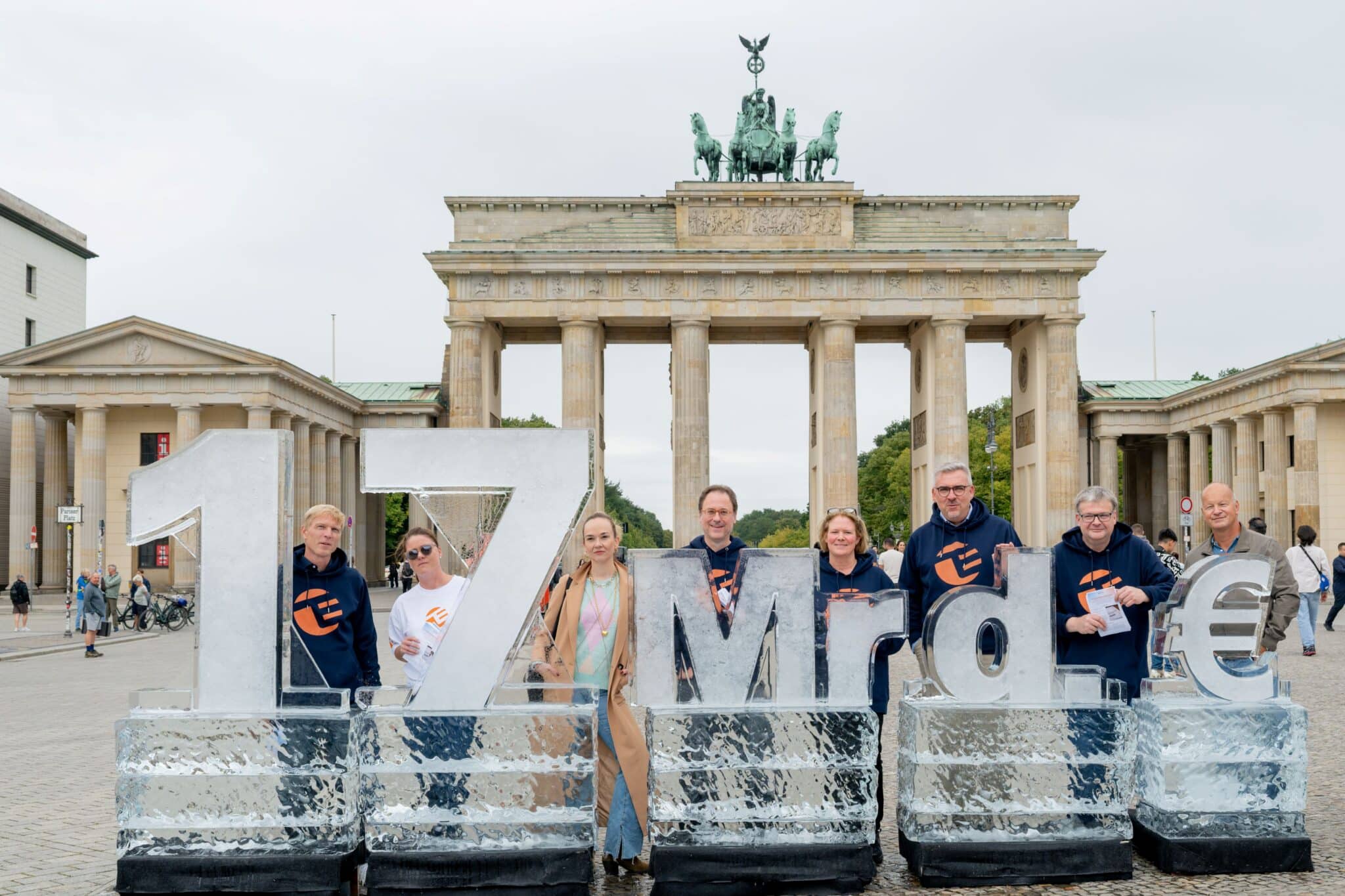 Gruppe von Menschen vor dem Brandenburger Tor, posierend mit einer großen Eisskulptur, die "17 Mrd. €" zeigt.