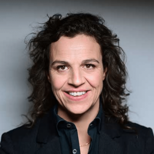 Porträt einer lächelnden Frau mit lockigem Haar, die einen schwarzen Blazer trägt, vor einem neutralen Hintergrund.