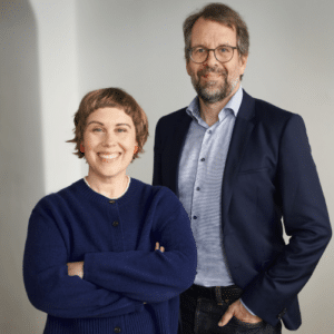 Zwei Personen stehen lächelnd nebeneinander, eine Frau mit kurzen Haaren in einem blauen Pullover und ein Mann in einem A...