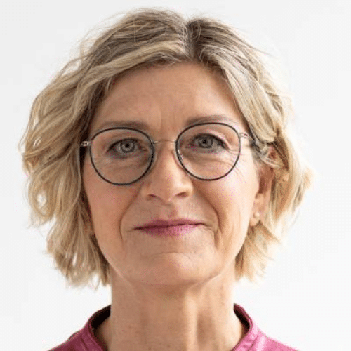 Porträt einer Frau mit blonden, lockigen Haaren und Brille, die in die Kamera lächelt. Sie trägt ein pinkes Oberteil.