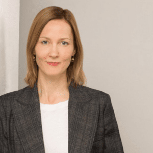 Frau mit blonden Haaren in einem grauen Blazer und weißem Shirt, vor neutralem Hintergrund.