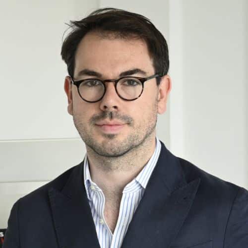 Porträt eines Mannes mit Brille, dunklem Anzug und gestreifter Hemd, vor einem hellen Hintergrund.