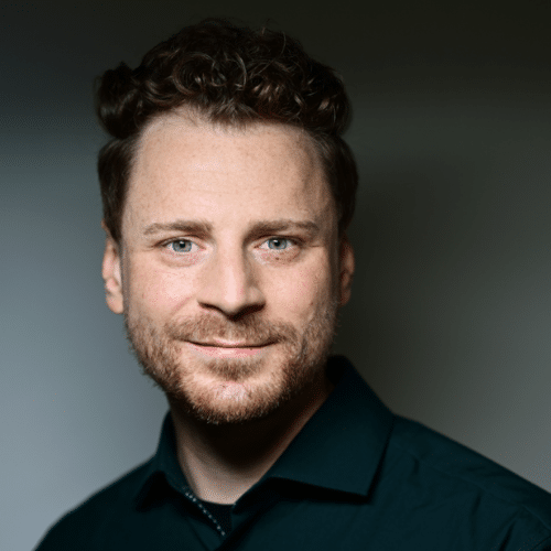 Porträt eines Mannes mit lockigem Haar und Bart, der in die Kamera lächelt, vor einem neutralen Hintergrund.