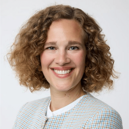 Porträt einer lächelnden Frau mit lockigem, blondem Haar, die einen hellen Blazer trägt.