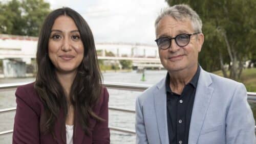 Zwei Personen sitzen am Ufer eines Gewässers, umgeben von Bäumen und einer Brücke im Hintergrund.